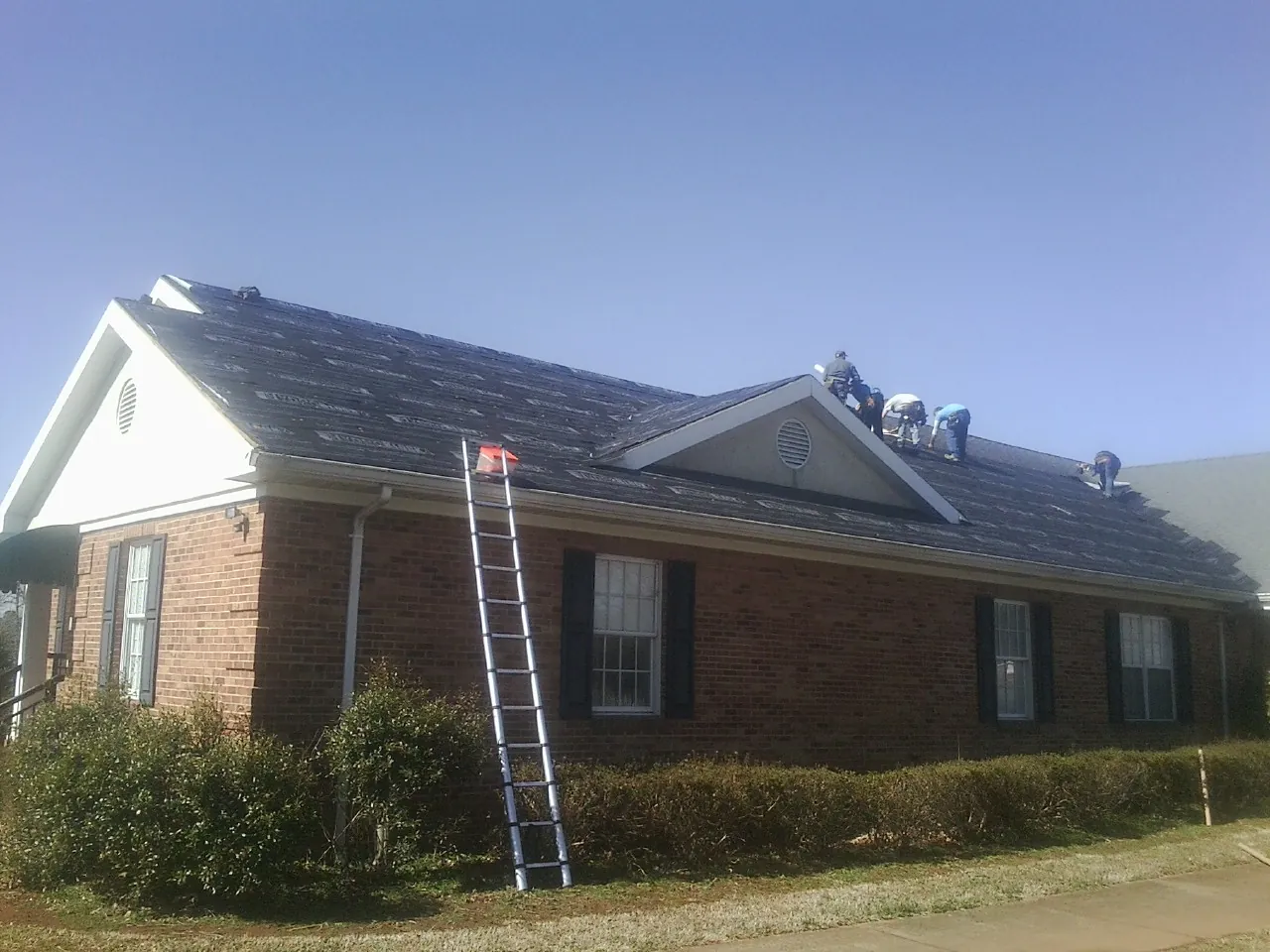 Crew tearing shingles off a brick ranch roof with a ladder set — Emergency Roof Repair work by our crew in New Port Richey